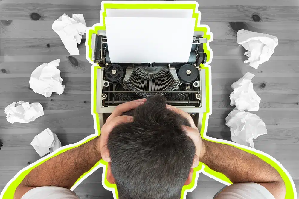 a top view of a man with his head in his hands looking at a typewriter with crumpled papers on a desk beside him