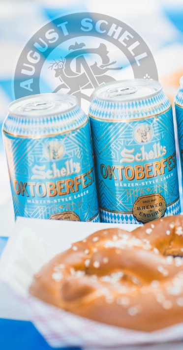 cans of schells oktoberfest beer next to a pretzel with the schell's watermark seal in the background